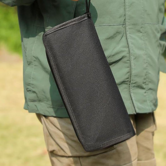 A person holding a black wallet with a green jacket and beige pants outdoors.